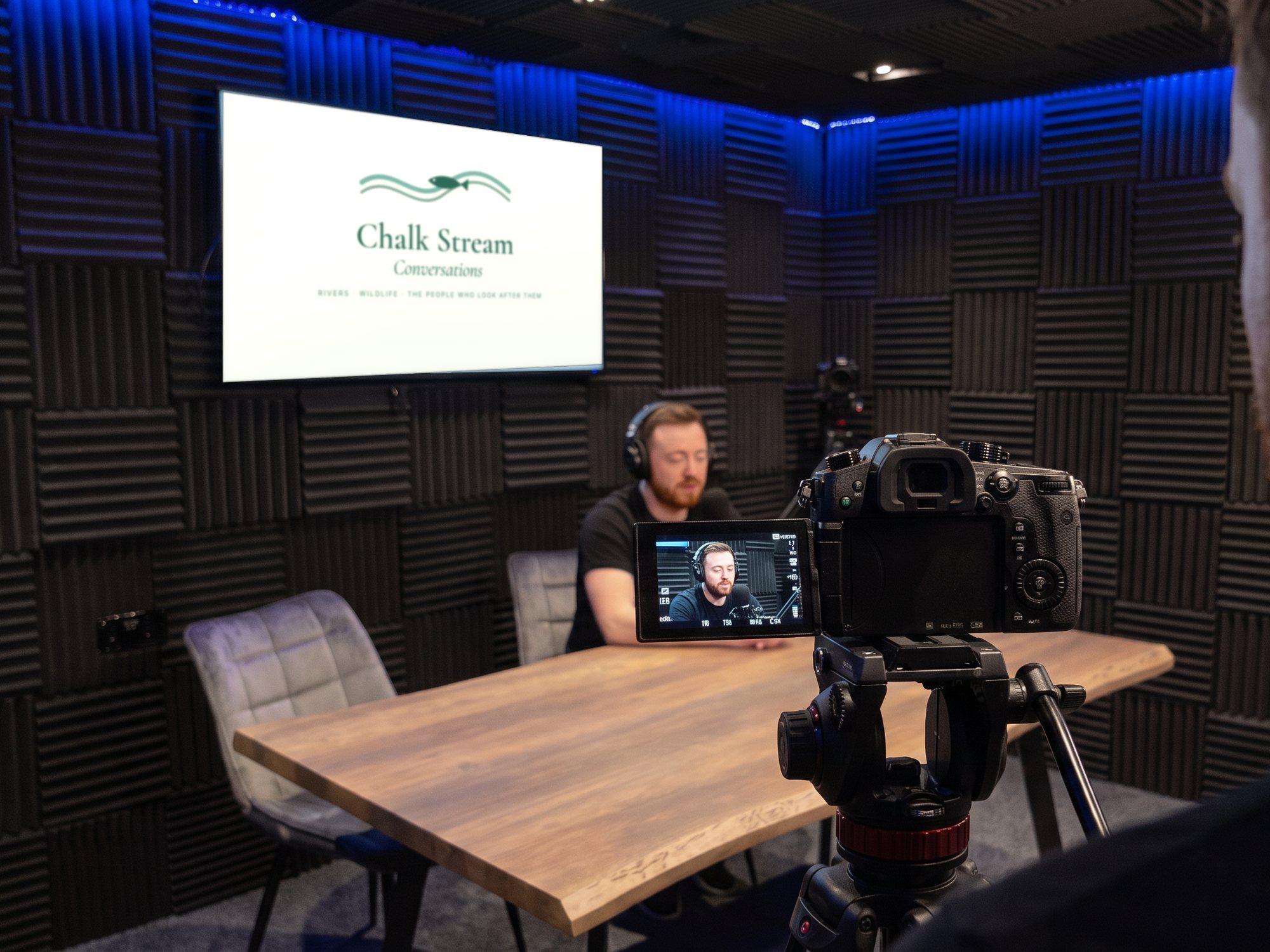 Over the camera operator's shoulder, a presenter at the table with the show card on the screen