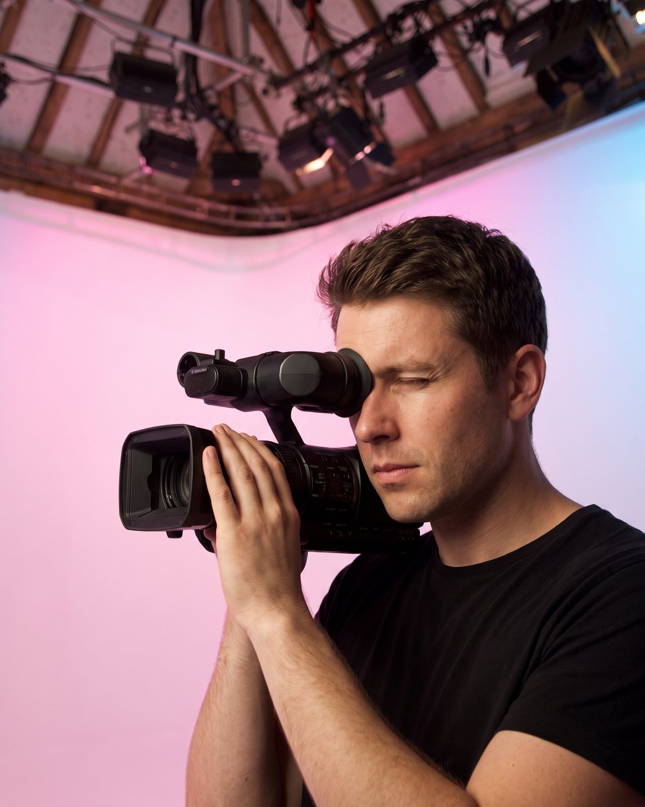 A camera operator at the viewfinder of a shoulder camcorder, the cove glowing pink behind