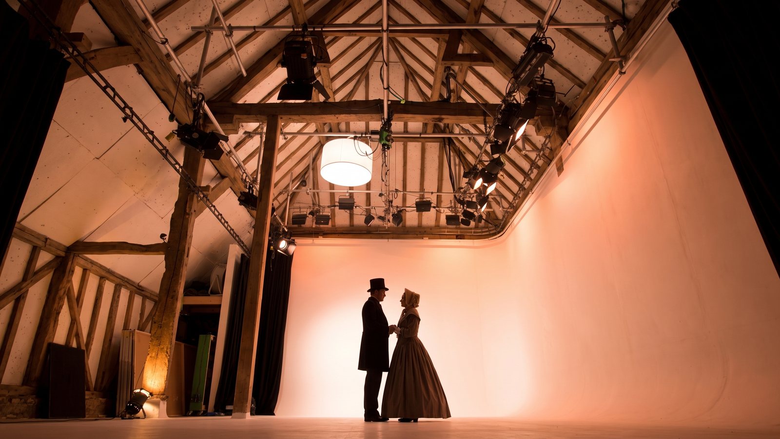 Two actors in Victorian costume against the cove, lit amber under the oak roof