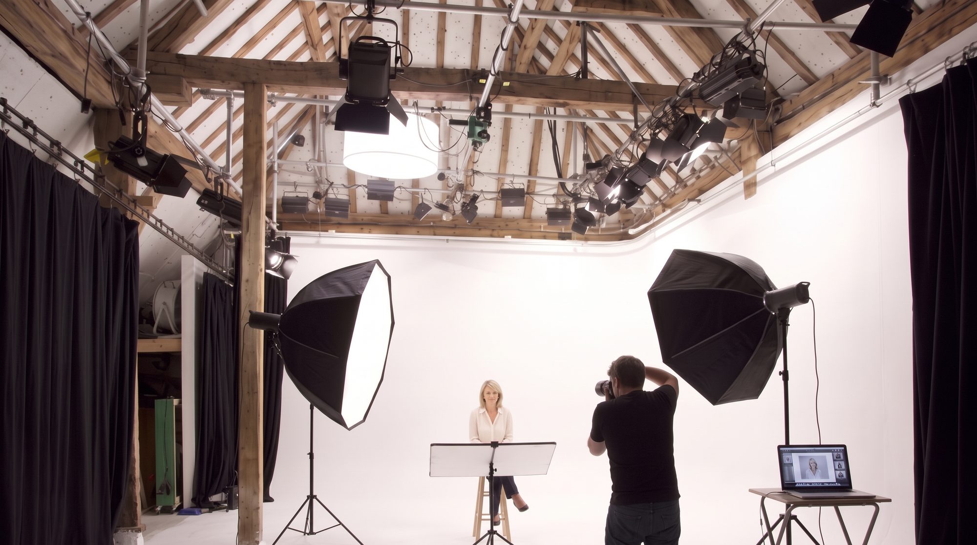 A businesswoman on a stool against the white cove while the photographer shoots, the oak roof and lighting grid above