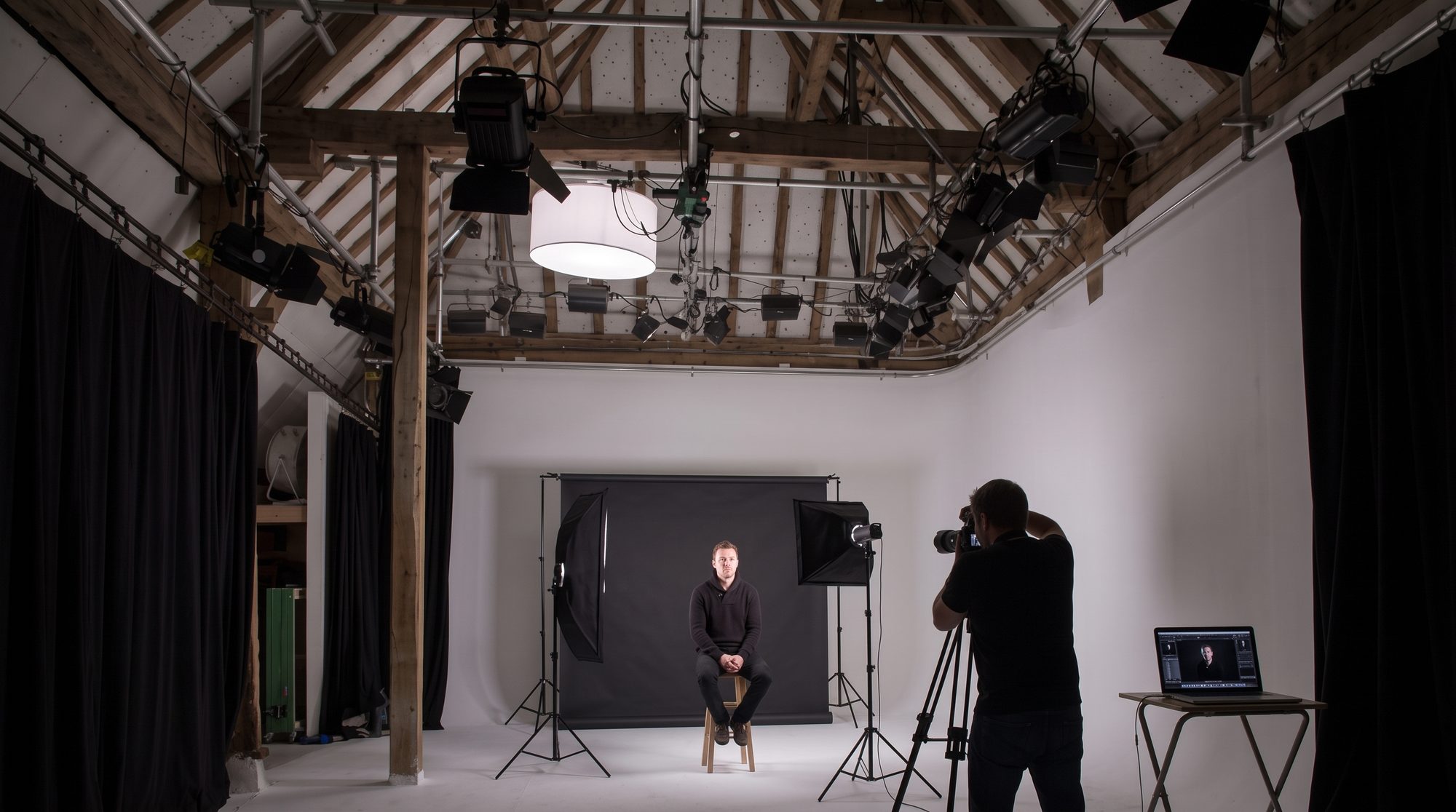 A man on a stool against a black backdrop, lit by one softbox and a strip light